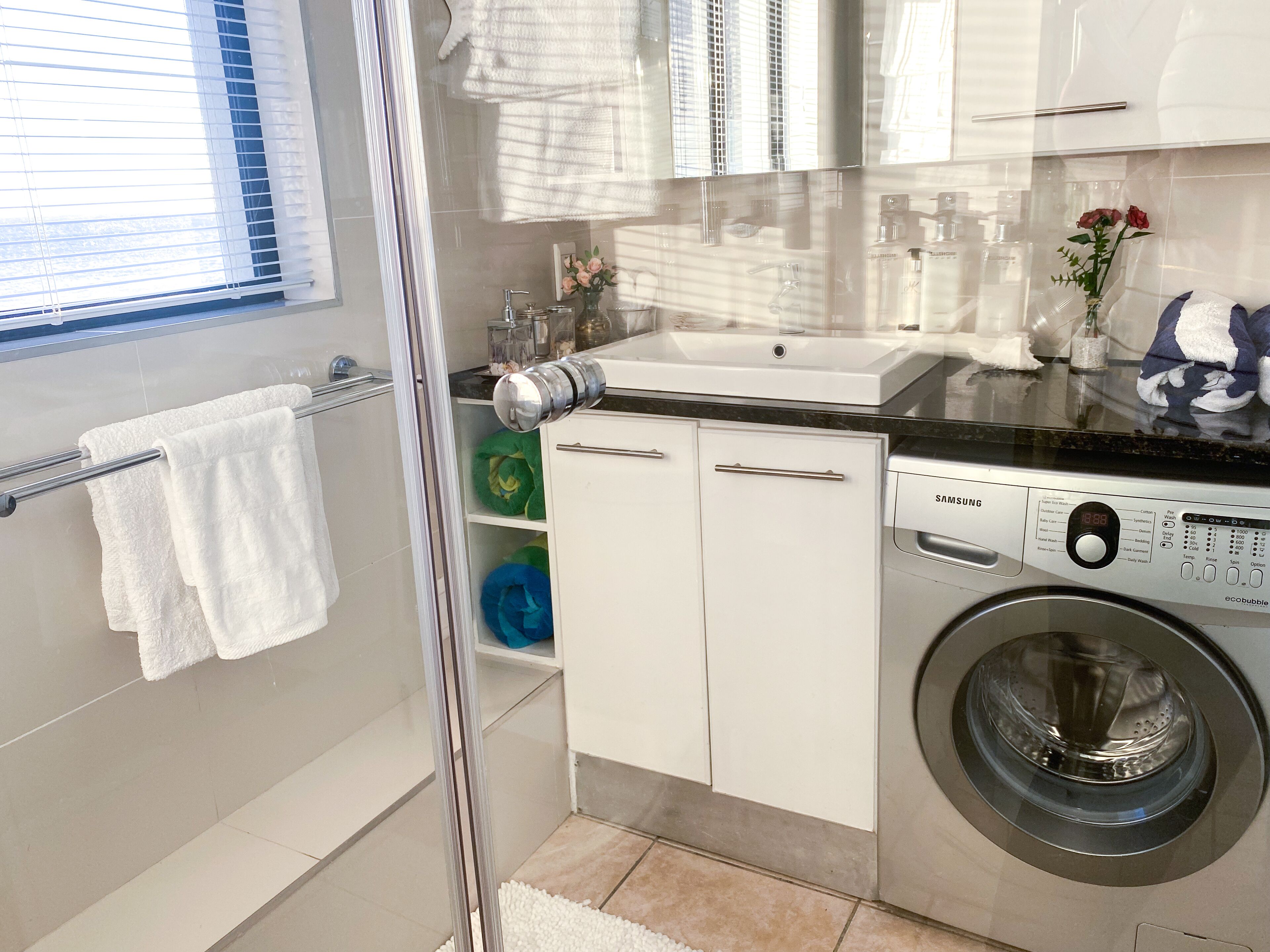 A modern bathroom with a glass shower enclosure, a built-in sink, a washing machine, and a towel rack.