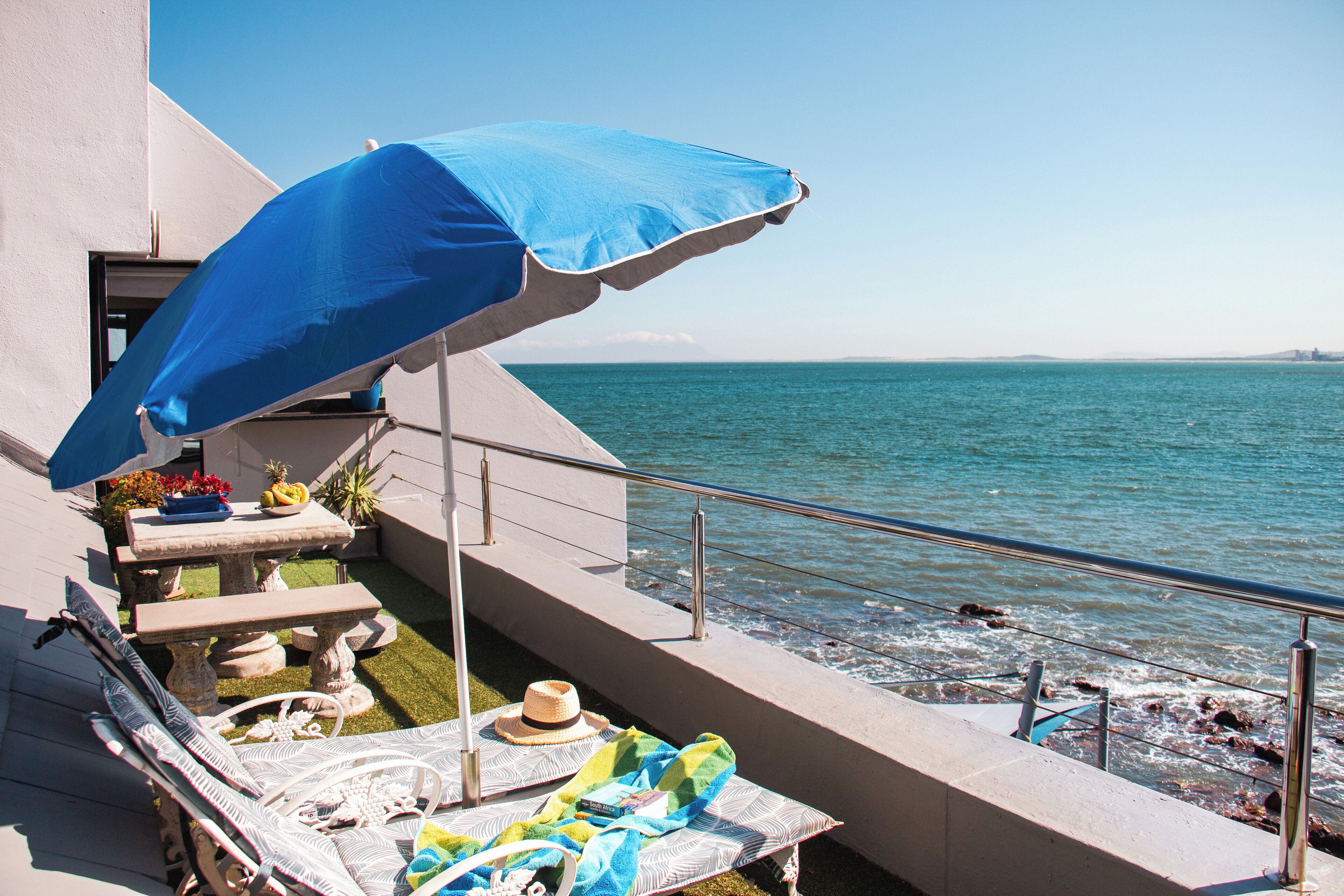 A balcony with a blue umbrella, a table with flowers, and a view of the sea.