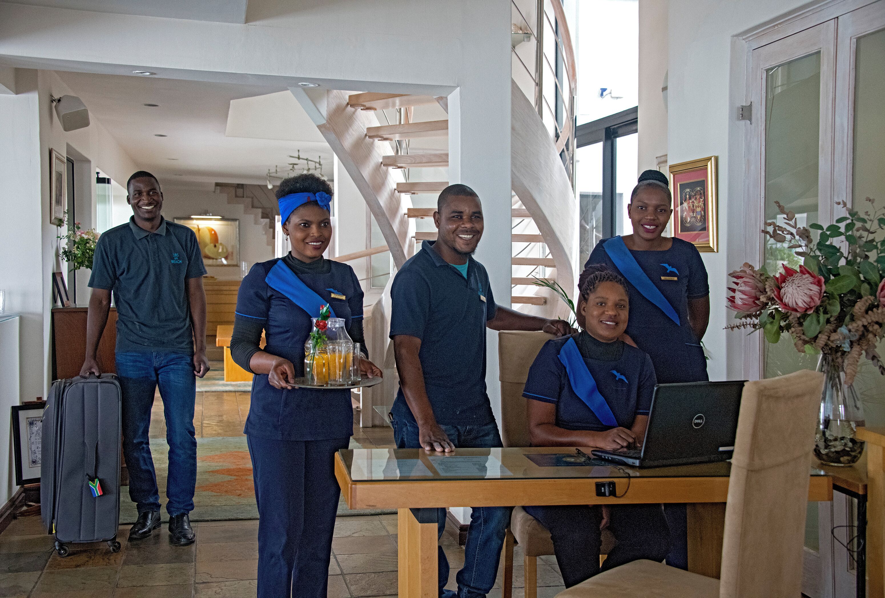 A group of people in a hotel lobby with a staircase, a table with a laptop, and a tray with drinks.