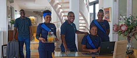 A group of people in a hotel lobby with a staircase, a table with a laptop, and a tray with drinks.
