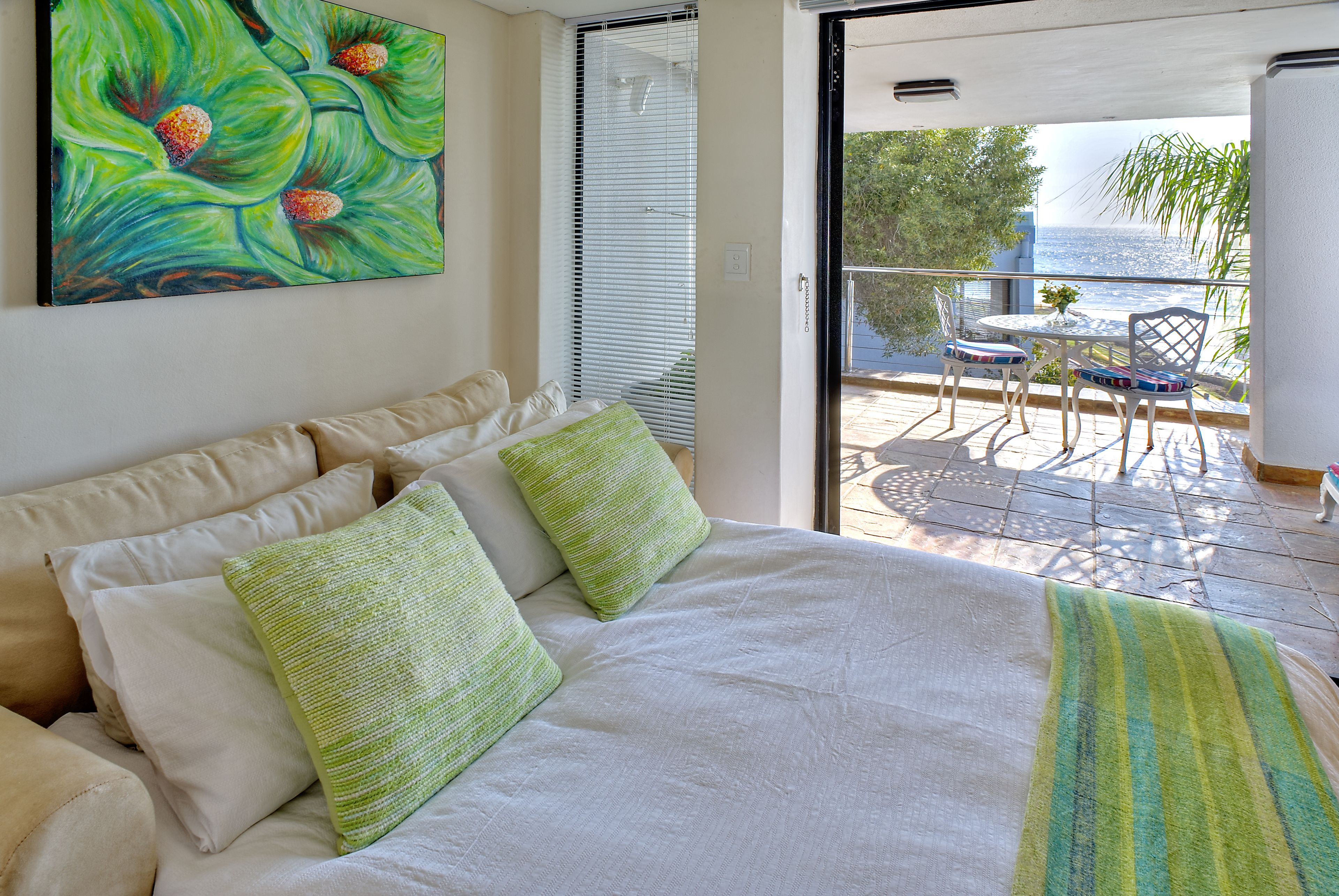 A bedroom with a bed, pillows, and a painting. There is a balcony with a table and chairs.