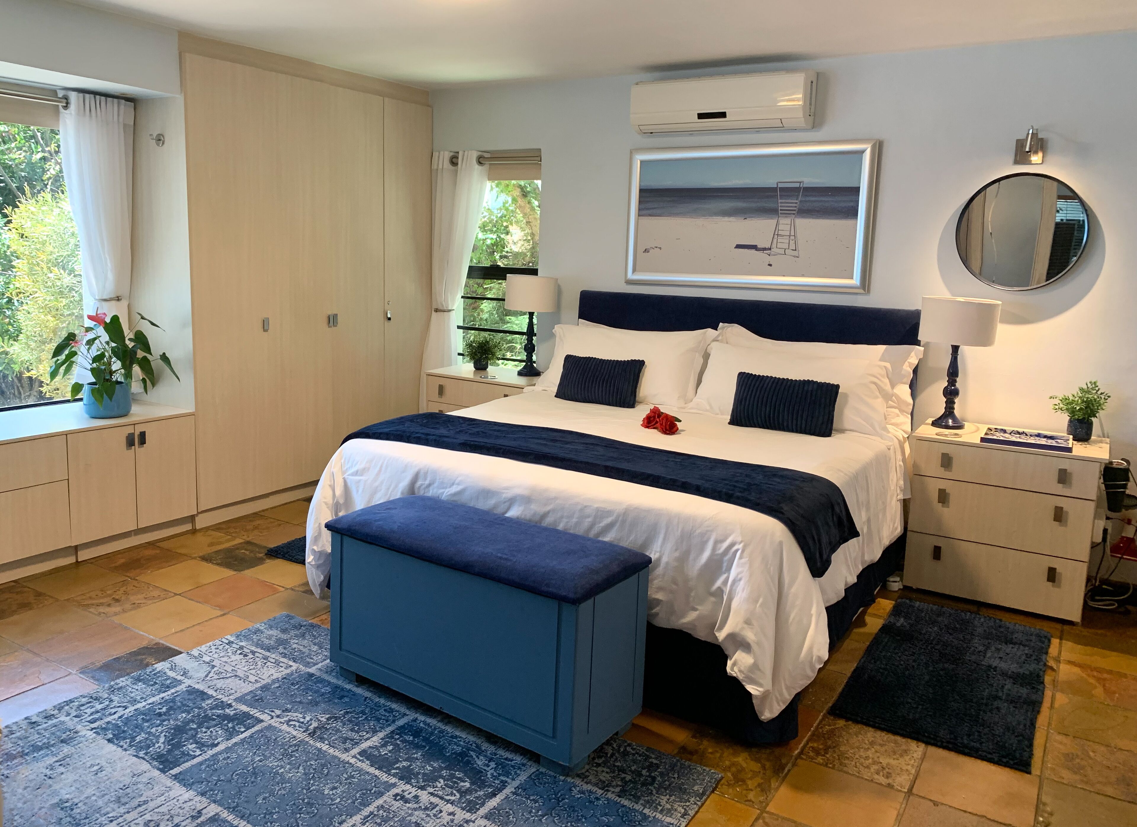 Une chambre moderne avec un grand lit, des tables de chevet, une commode, une fenêtre avec des rideaux, une plante en pot et un climatiseur fixé au mur.