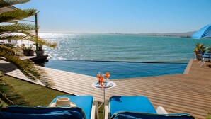 A wooden deck with lounge chairs and a table with drinks, overlooking a pool and the sea. - 185 on Beach Boutique Suites and Apartments (Cape Town)