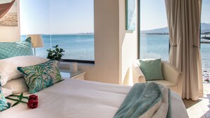 A bedroom with a large window offering a view of the sea and a distant city skyline. - 185 on Beach Boutique Suites and Apartments (Cape Town)