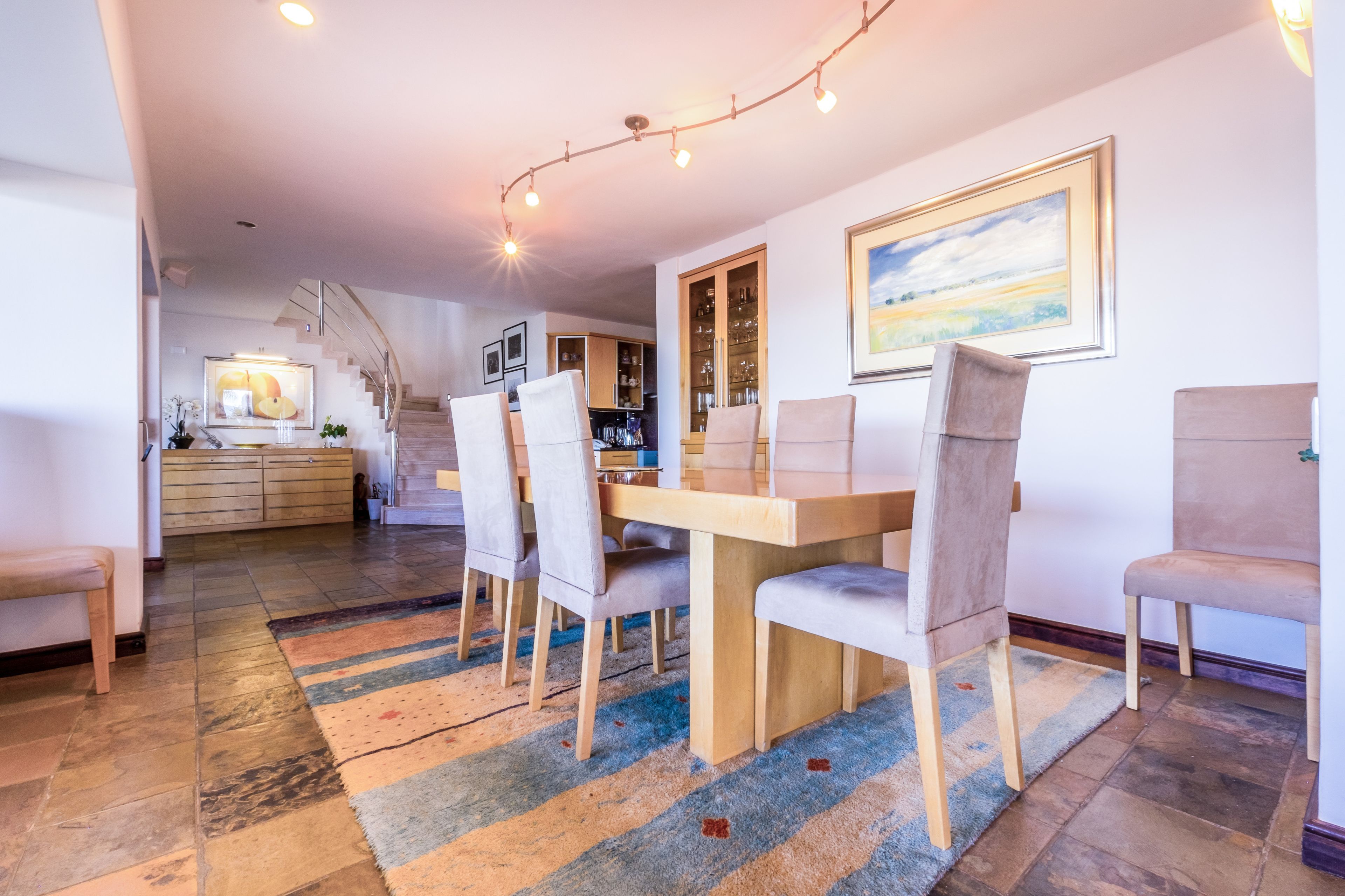 A dining area with a wooden table and chairs, a colorful rug, and a kitchen in the background.