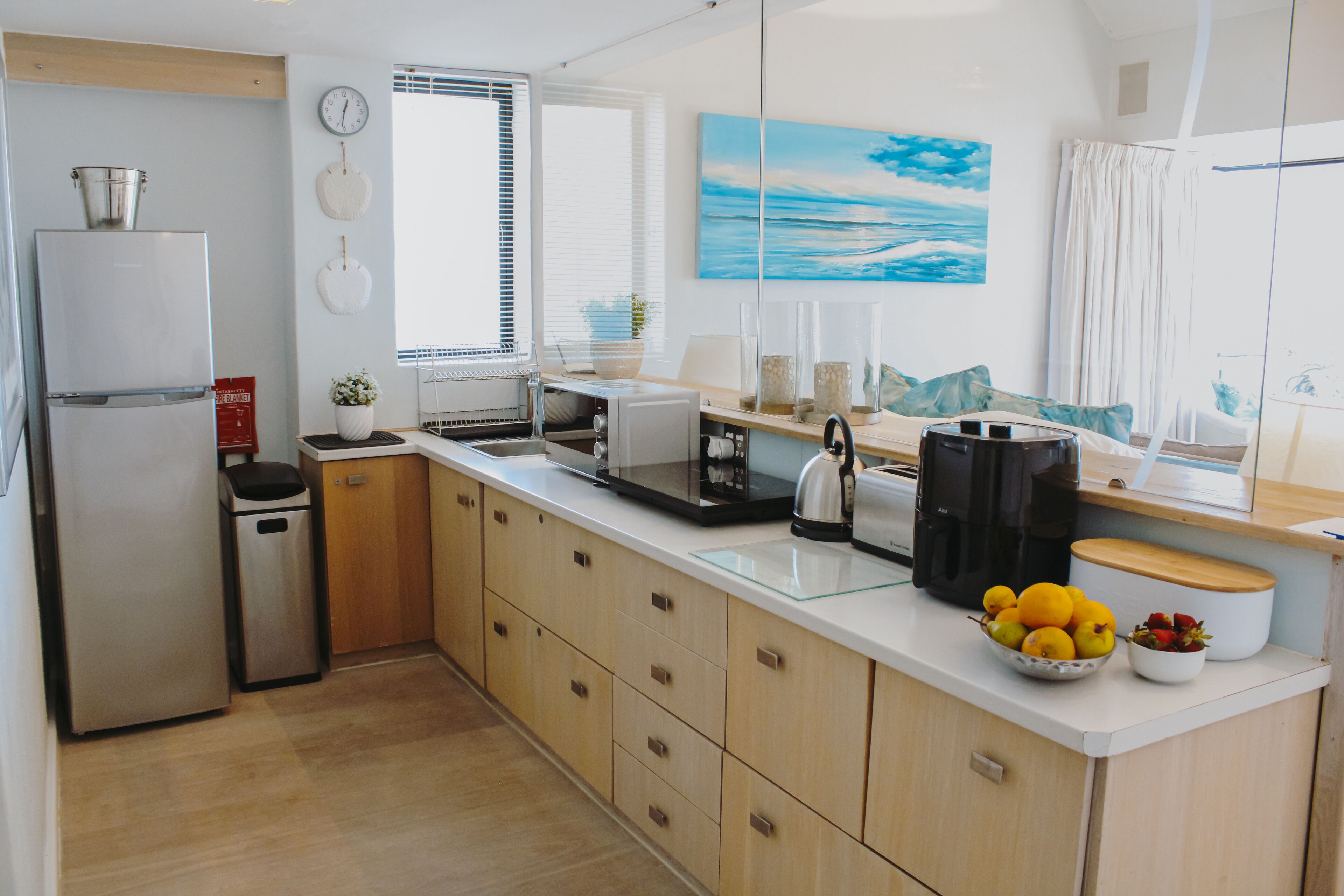 A modern kitchen with a refrigerator, sink, and coffee maker. There is a window with blinds and a painting on the wall.
