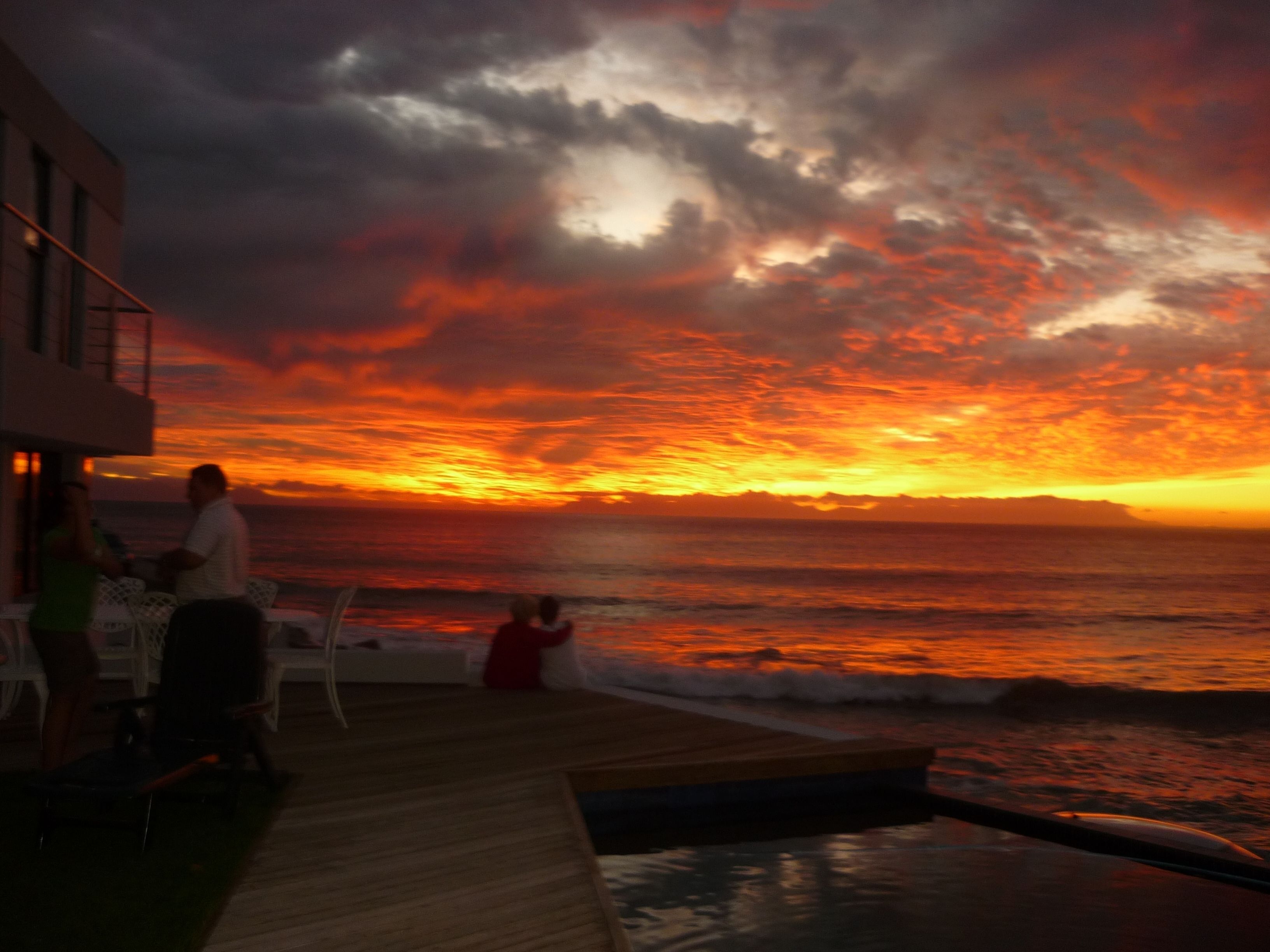 Solnedgangsudsigte fra en træterrasse med udsigt over havet, inklusive en pool og udendørs siddeområde.
