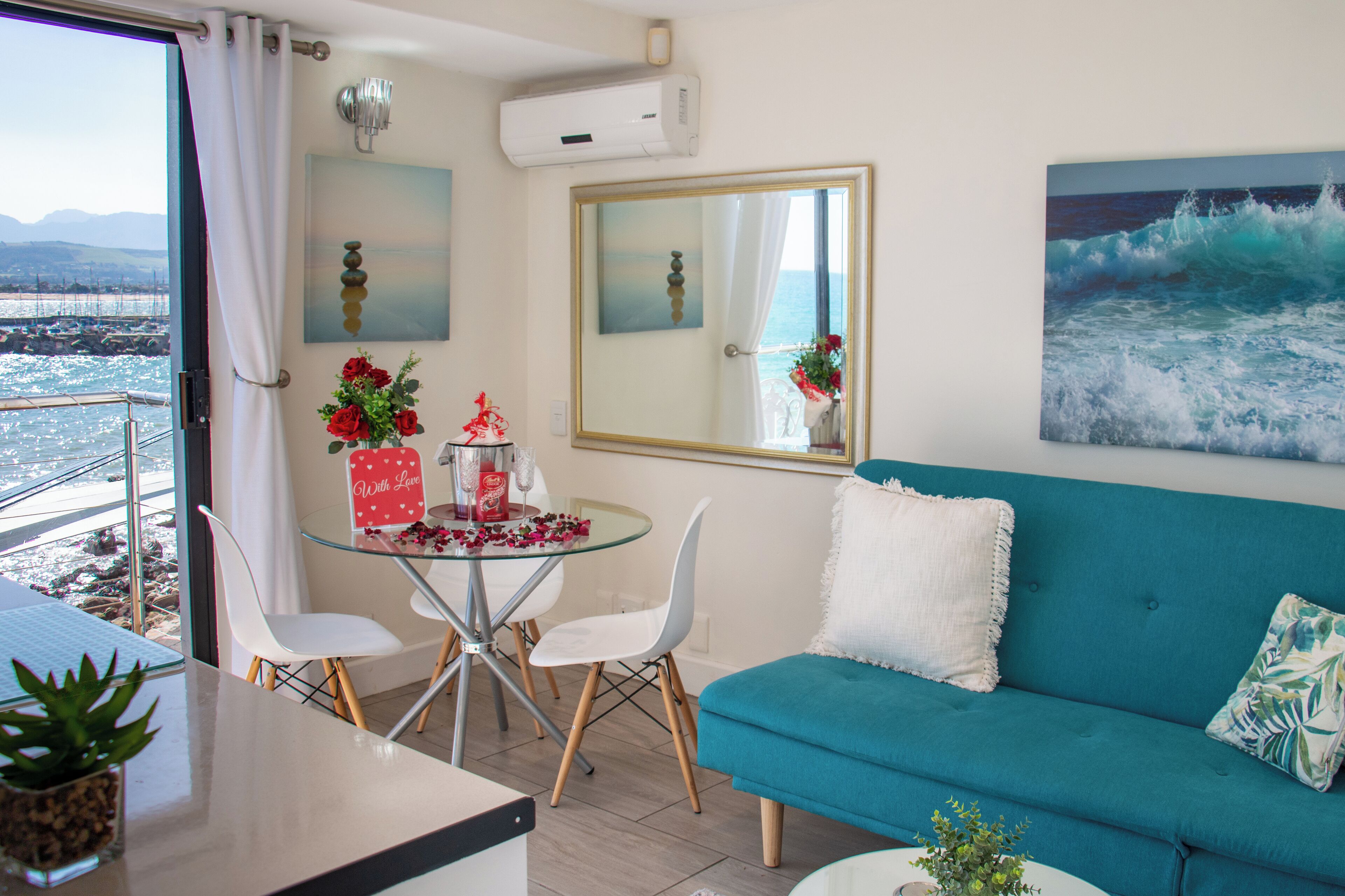 A room with a teal sofa, a glass table with chairs, a view of the sea through a window, and a painting of waves.