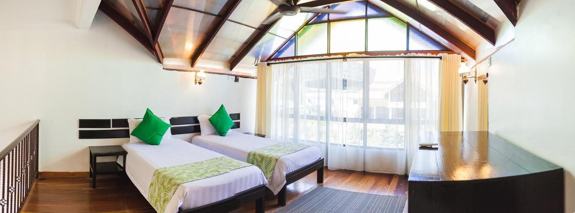 A room with two beds, a bench, a desk, and a large window with curtains.