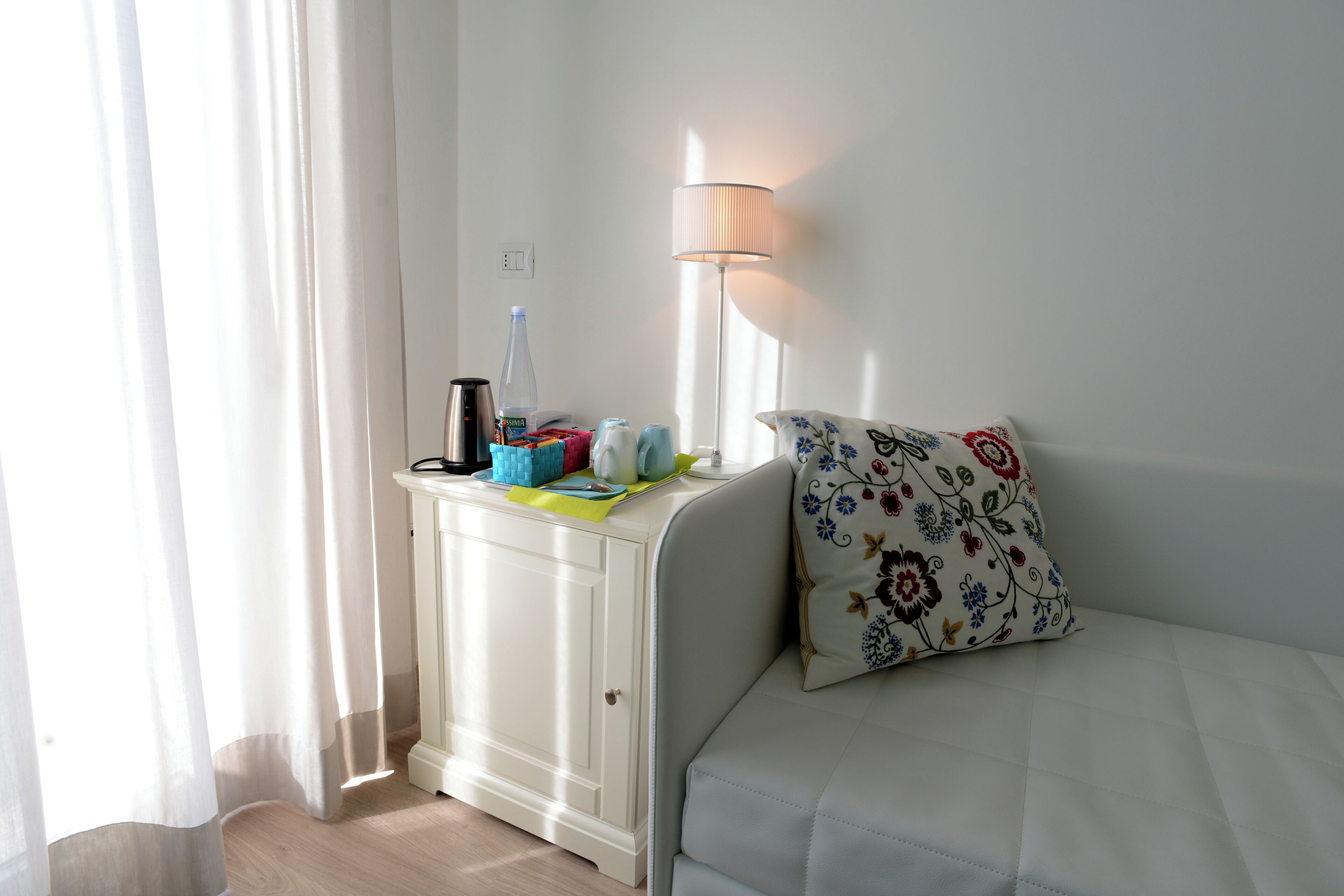 Una habitación con un sofá blanco, una mesita con una lámpara y una ventana con cortinas.