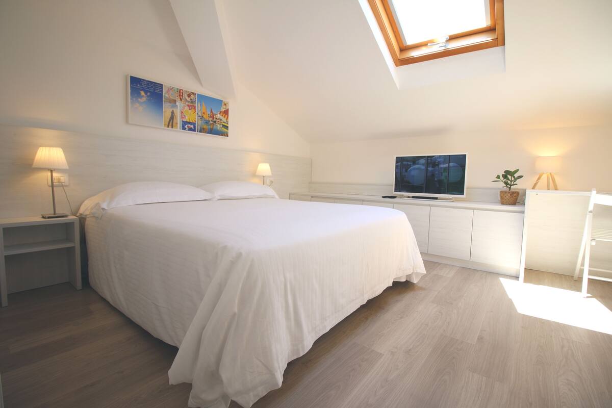 A bedroom with a bed, nightstands, a TV, a desk, and a skylight.