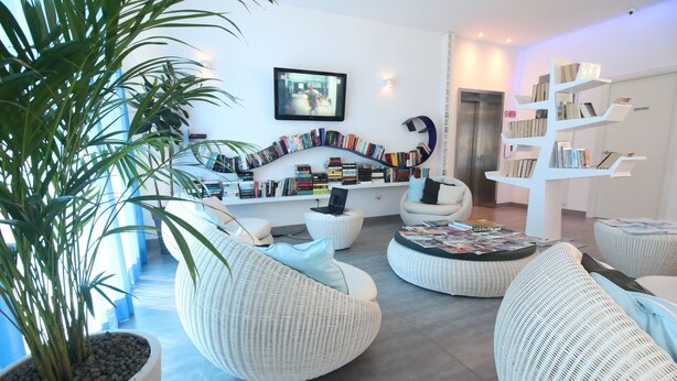 A modern living room with white wicker furniture, a bookshelf, and a television on the wall. - Maree Hotel (Cesenatico)