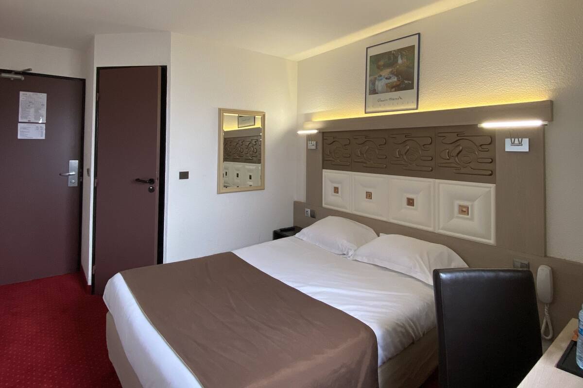 A hotel room with a bed, a desk, a chair, a telephone, and a door.