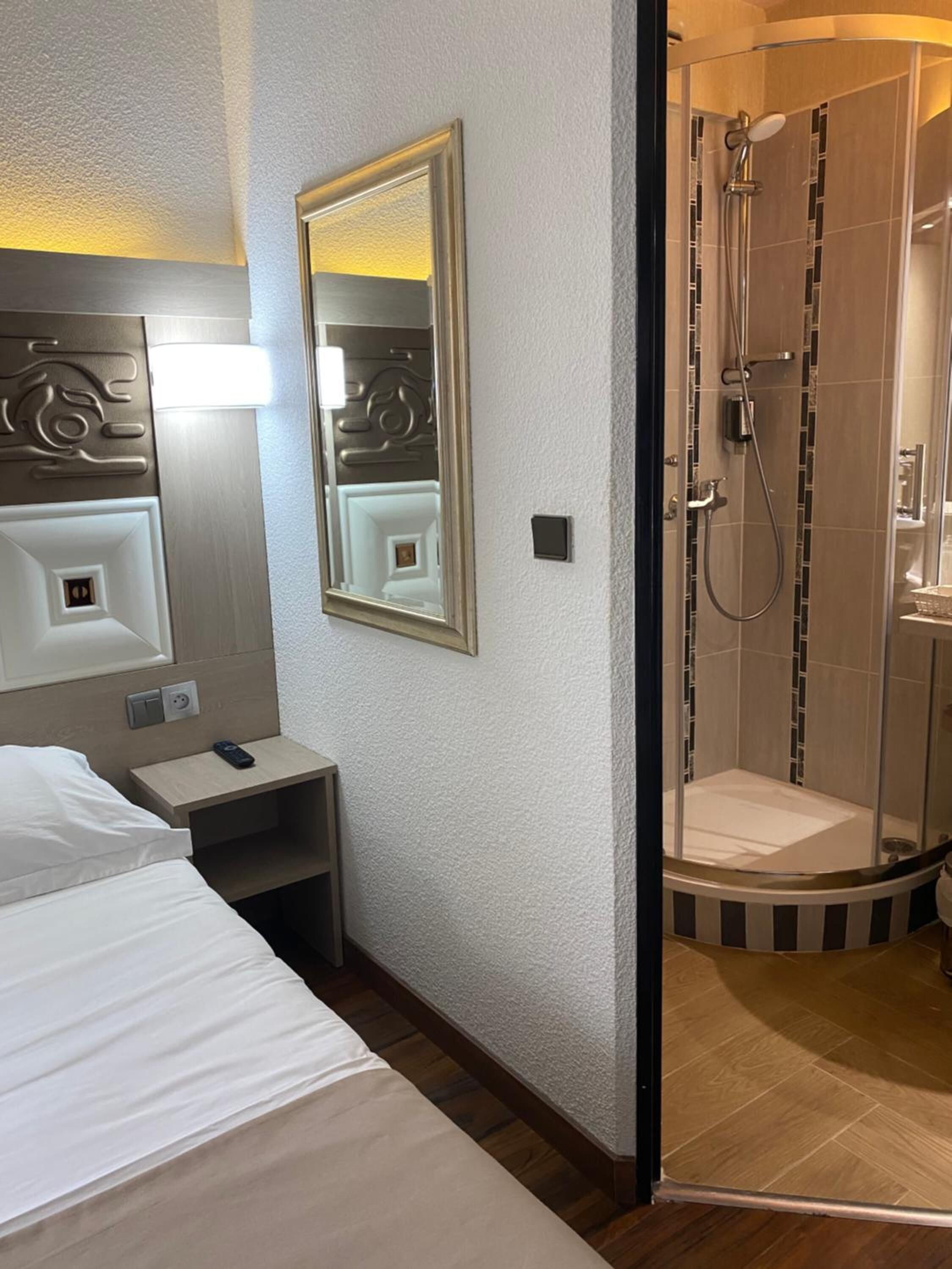 Una habitación de hotel con cama, mesita de noche, espejo y zona de ducha con puerta de cristal.