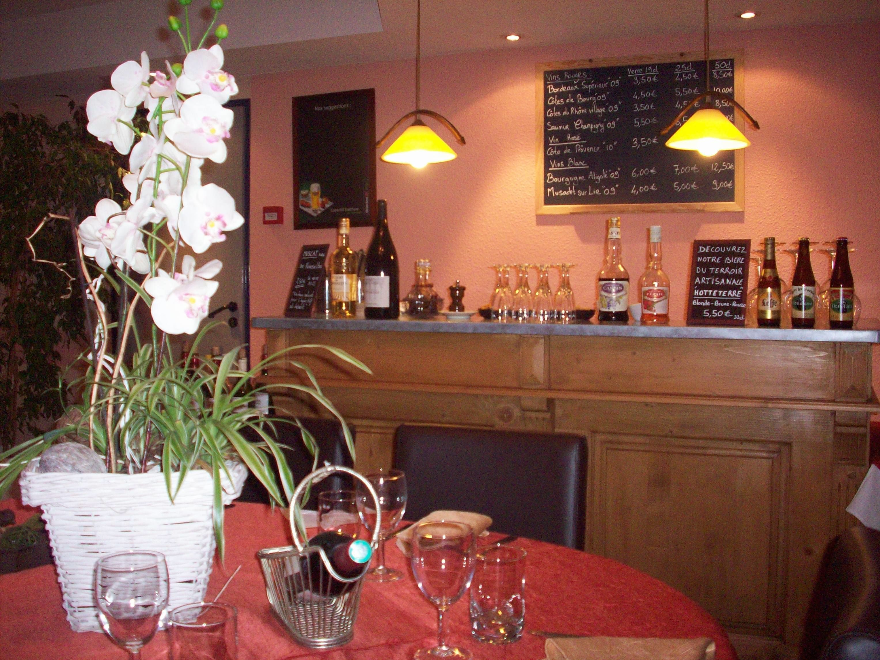 Interior de un restaurante con una pizarra de menú, botellas de vino, copas y una orquídea en maceta sobre la mesa.