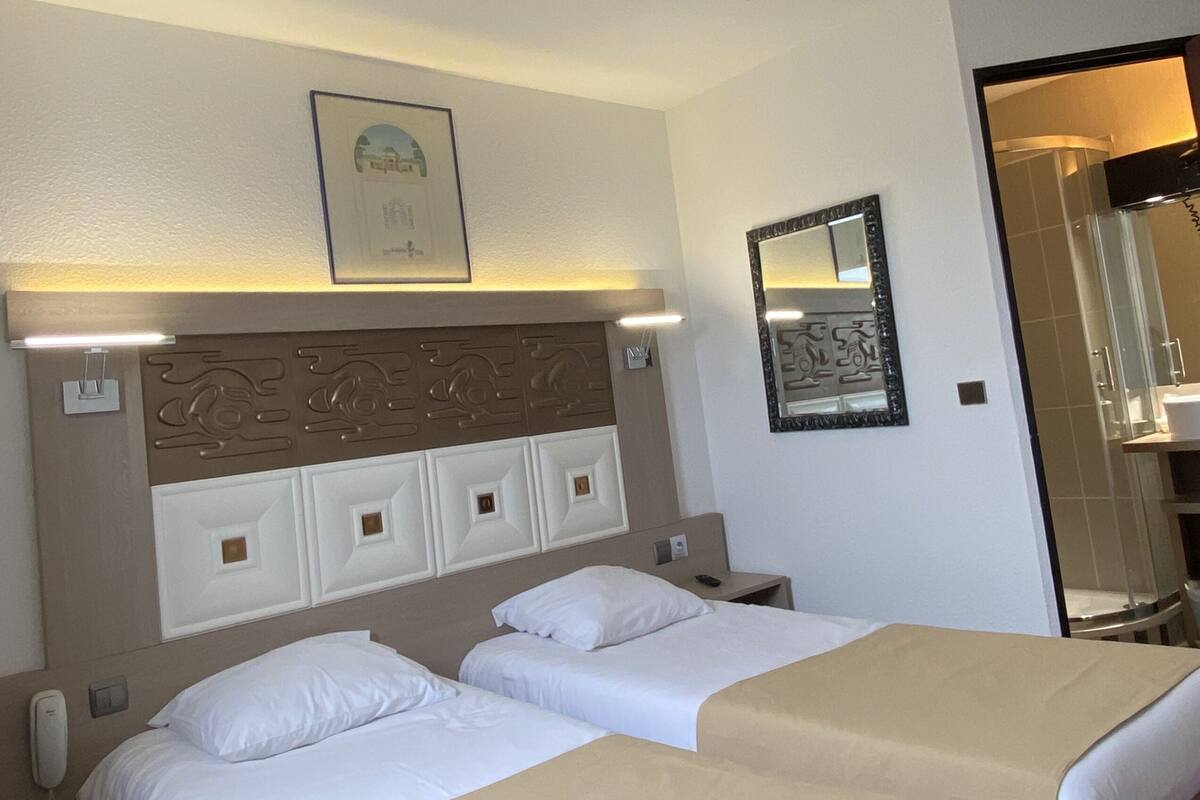 A hotel room with two beds, a headboard with decorative panels, a framed picture on the wall, a bedside table with a lamp, and a phone.