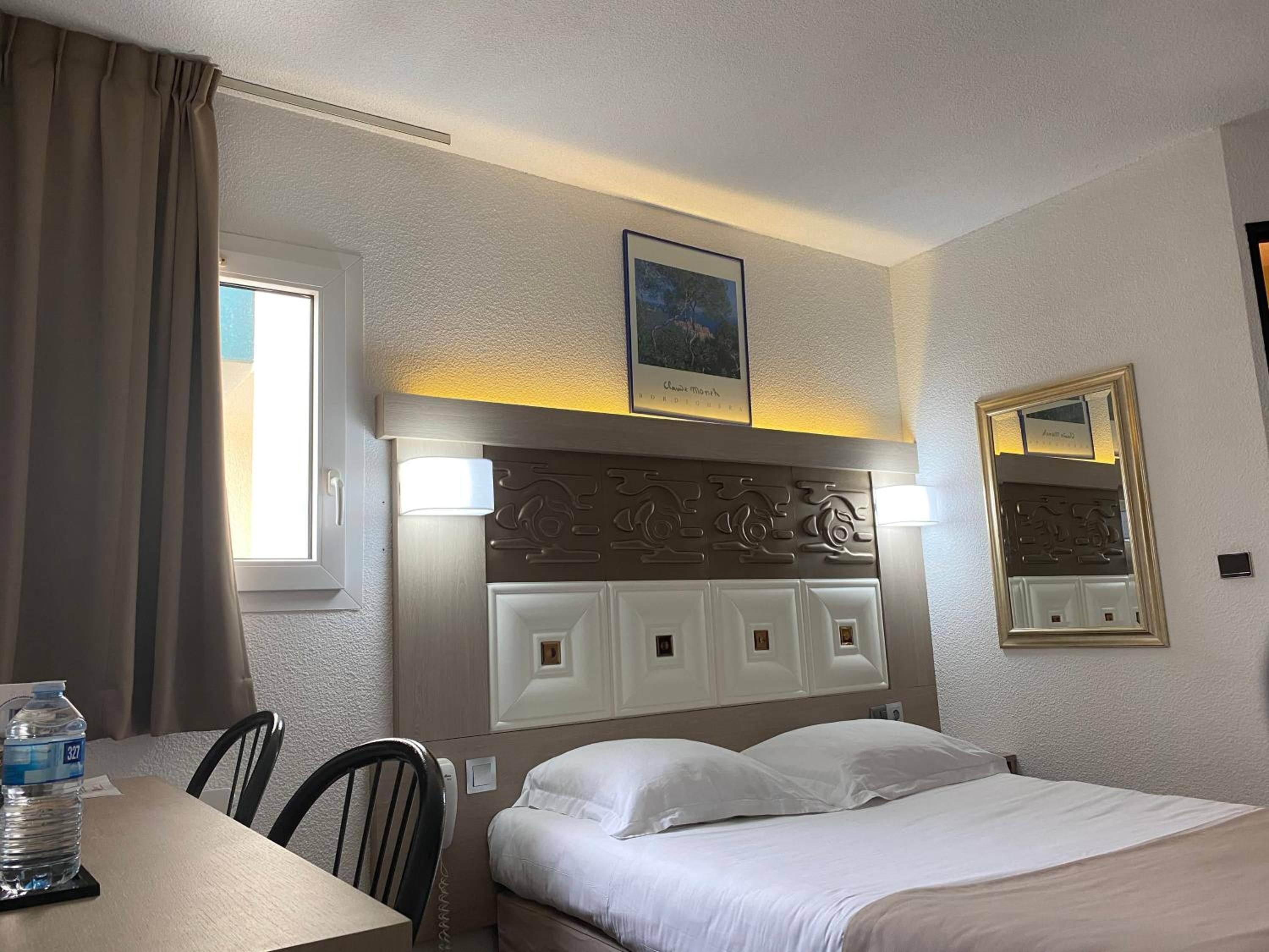 A hotel room with a bed, a table with a water bottle, a chair, a window with curtains, a wall-mounted picture, and a mirror.
