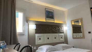 A hotel room with a bed, a table with a water bottle, a chair, a window with curtains, a wall-mounted picture, and a mirror.