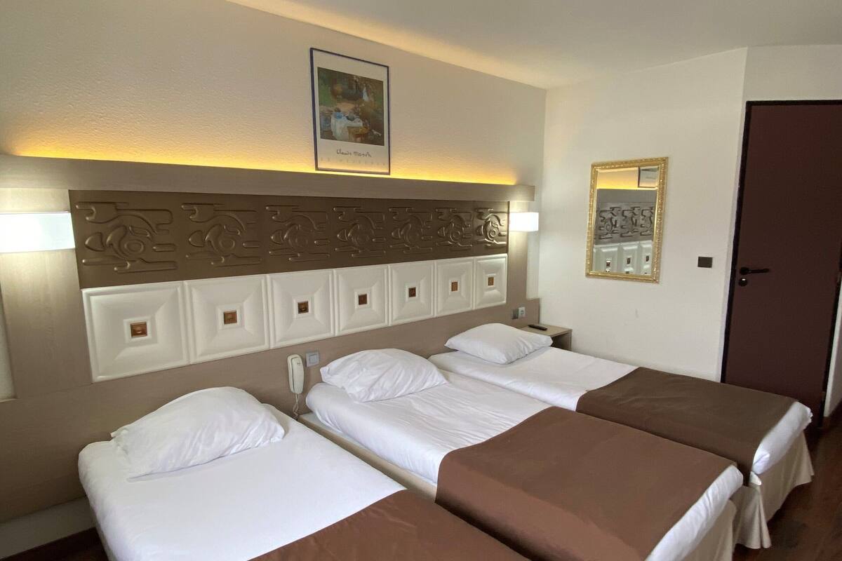 A hotel room with three single beds, a wooden floor, a headboard with decorative panels, a framed picture on the wall, and a door.