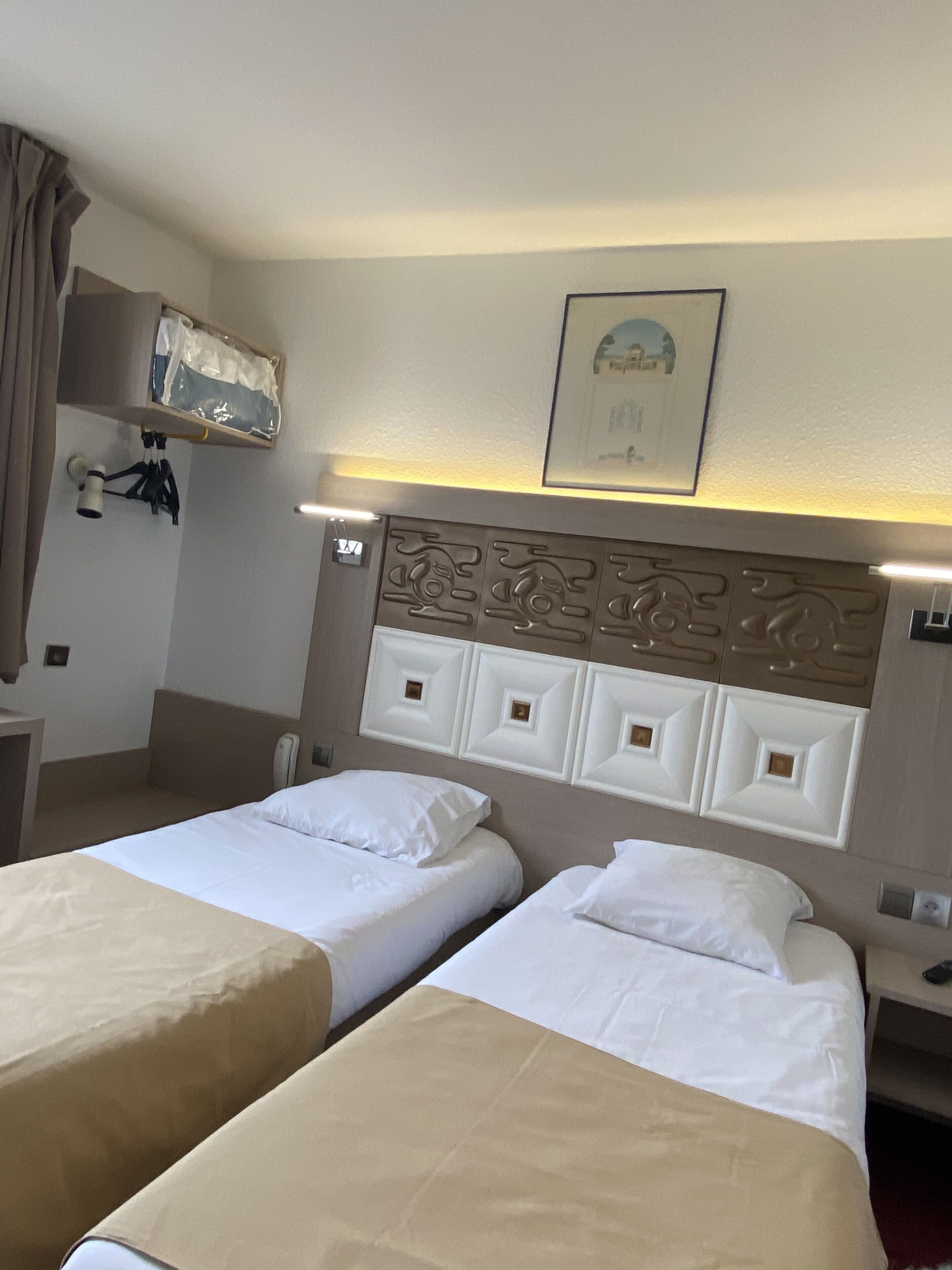 A hotel room with two beds, a headboard with decorative panels, a bedside table with a lamp, and a framed picture on the wall.