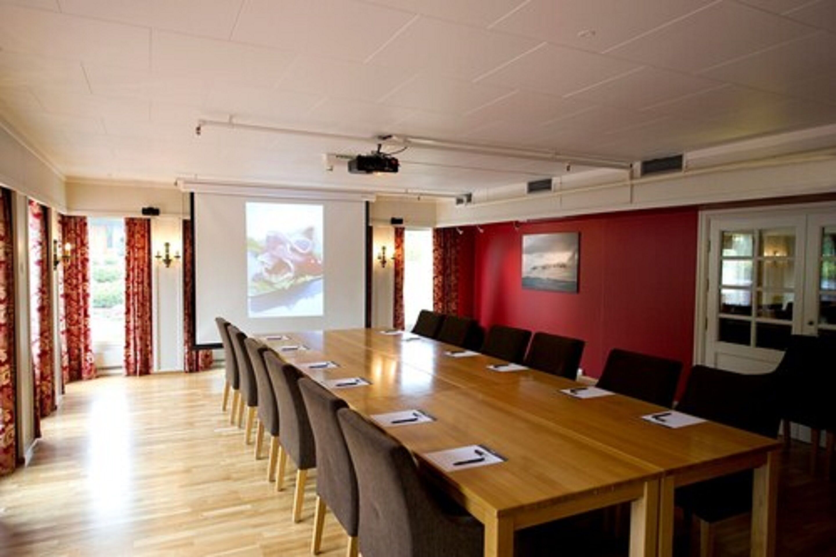 Un salón de conferencias con una larga mesa de madera, sillas, un proyector y una pared decorativa roja.