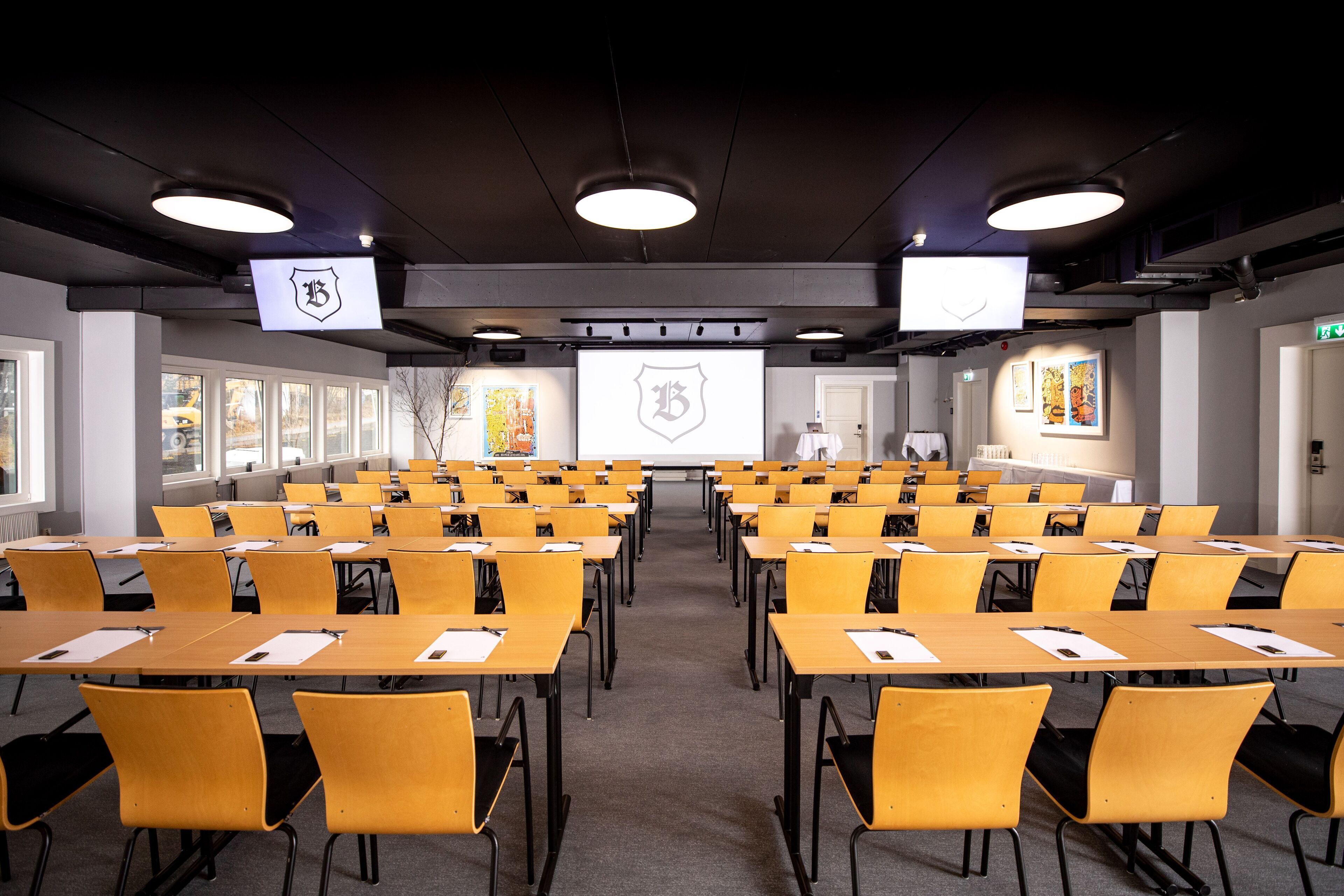 A conference room with rows of yellow chairs and tables, a projector screen, and a logo on the ceiling.
