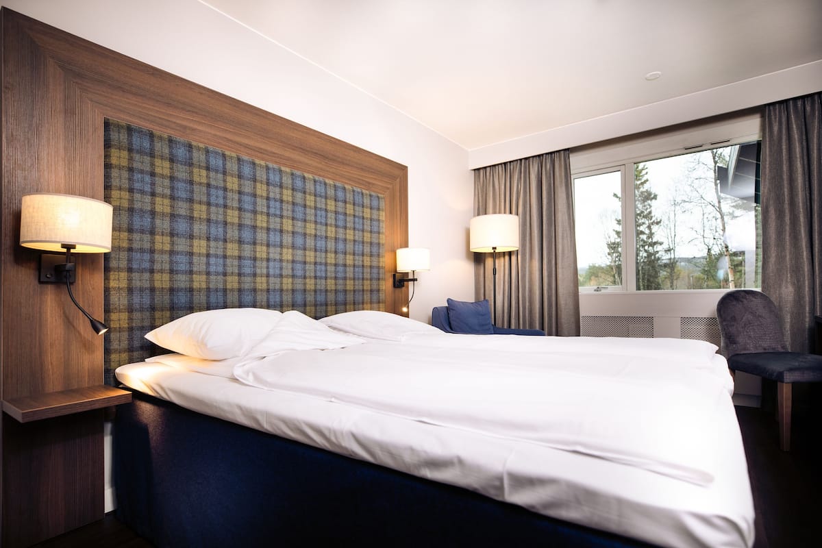 A hotel room with a large bed, two bedside lamps, a view of trees through the window, and a plaid-patterned headboard.