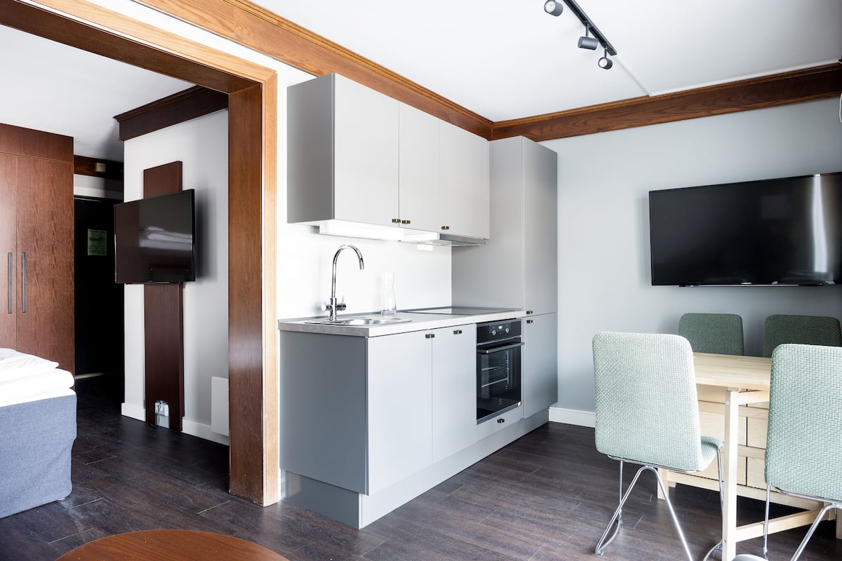A modern kitchen with white cabinets, a stainless steel range hood, and a built-in oven. There is a dining area with a wooden table and green chairs.