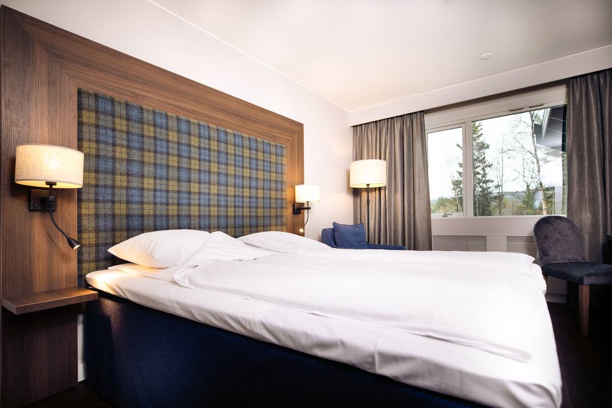 A hotel room with a large bed, two bedside lamps, a headboard with a plaid pattern, a chair, and a window with curtains.
