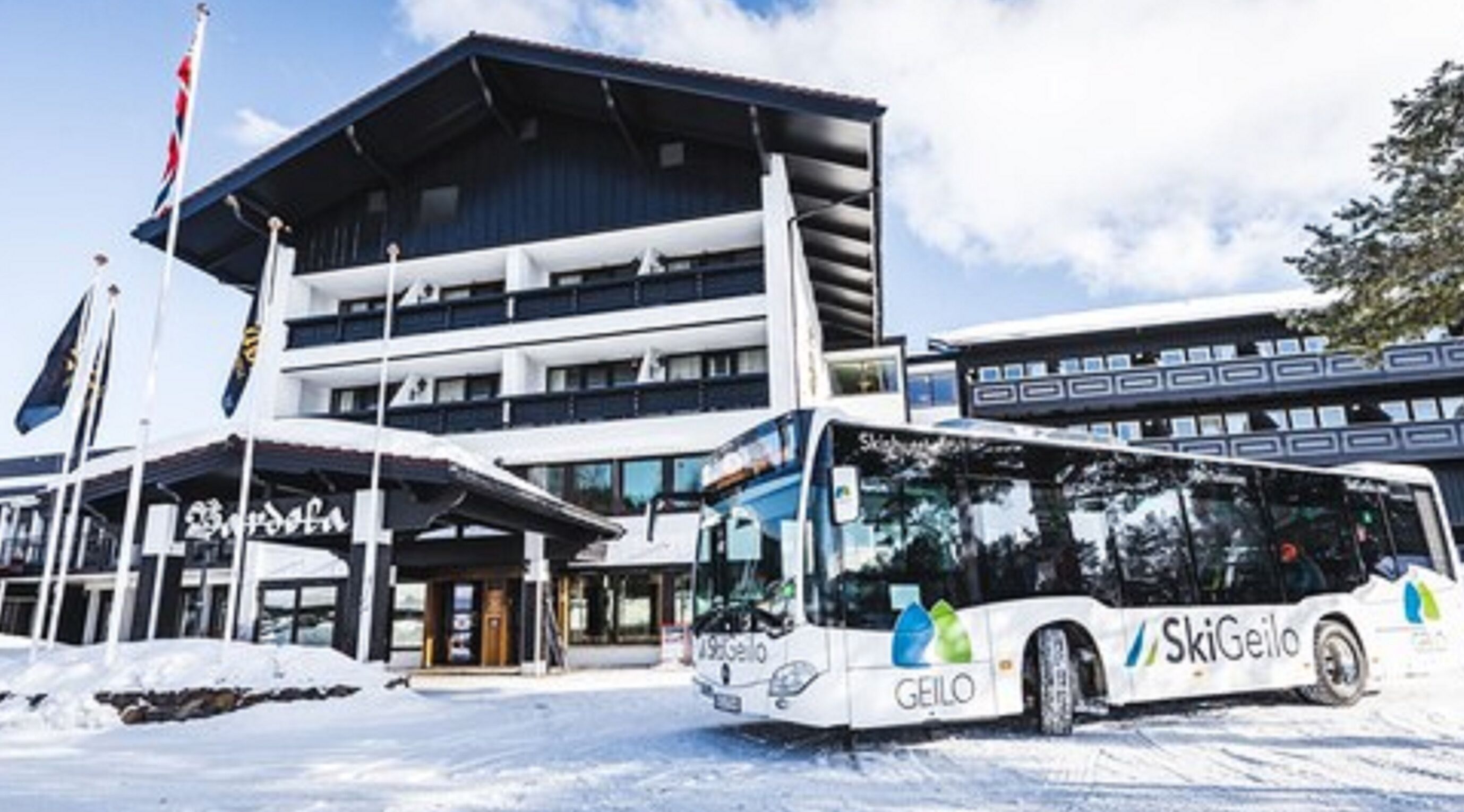 Ein Bus mit 'SkiGeilo'-Beschriftung vor einem mehrstöckigen Gebäude, im Hintergrund ist eine Seilbahn zu sehen.