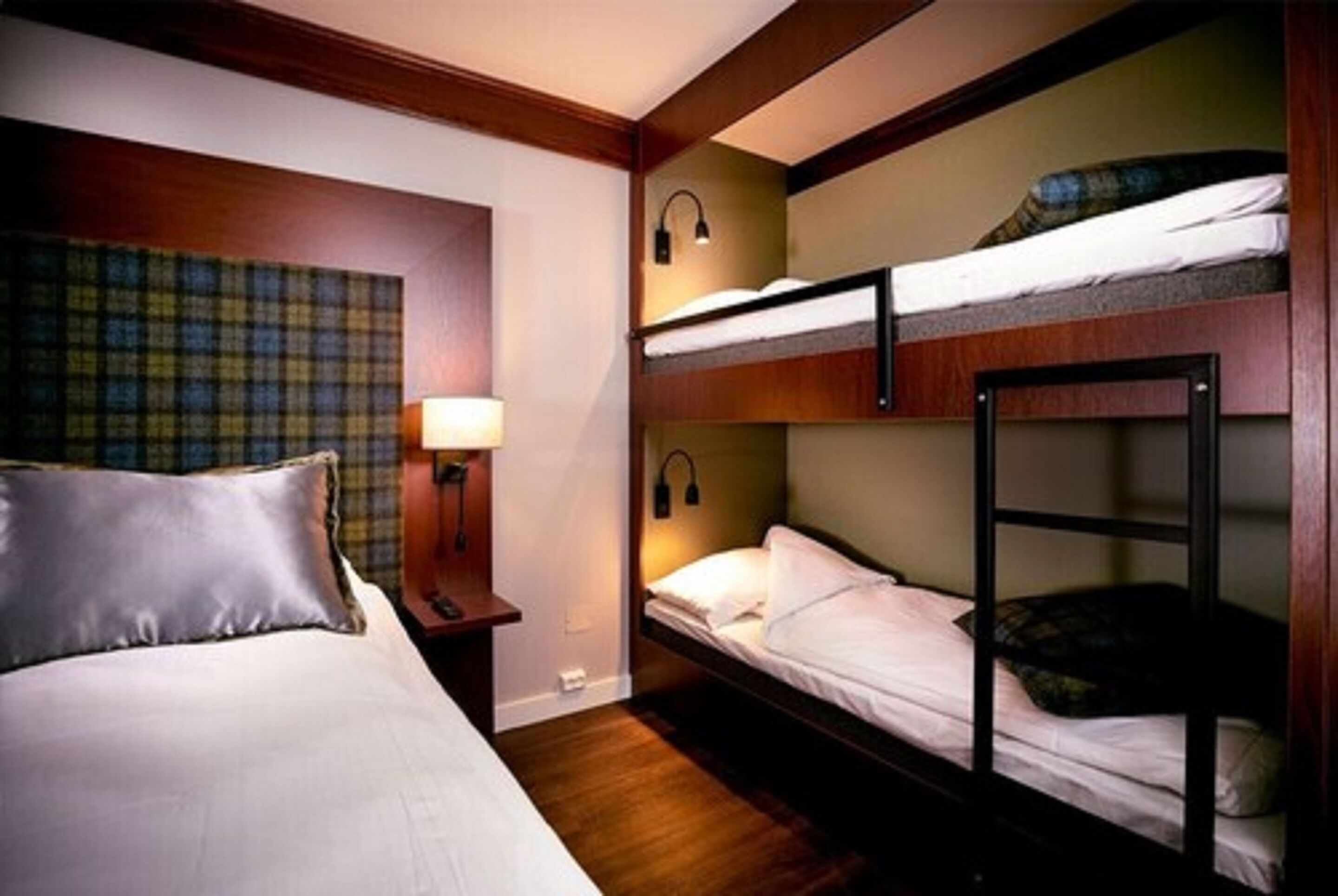 A hotel room with a single bed, a nightstand, a lamp, and two bunk beds.