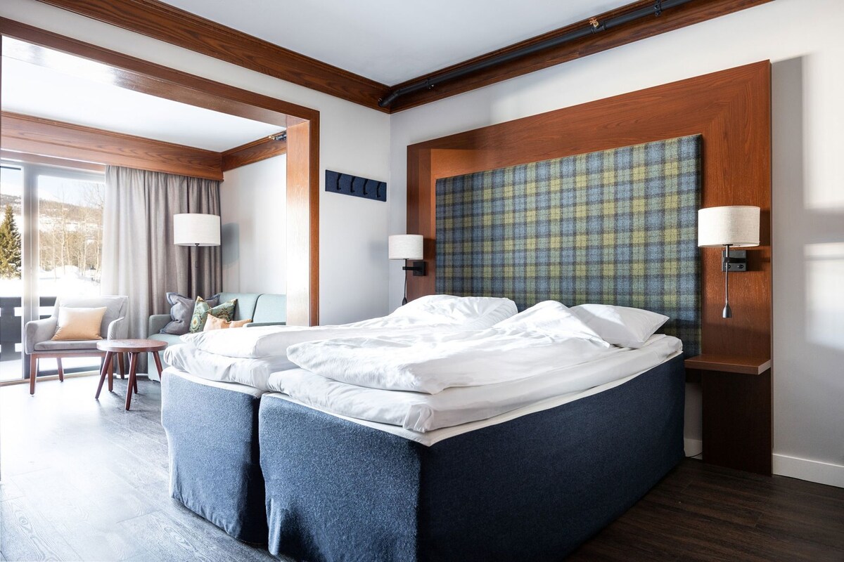 A hotel room with a large bed, a seating area with a chair and table, and a view through a window.