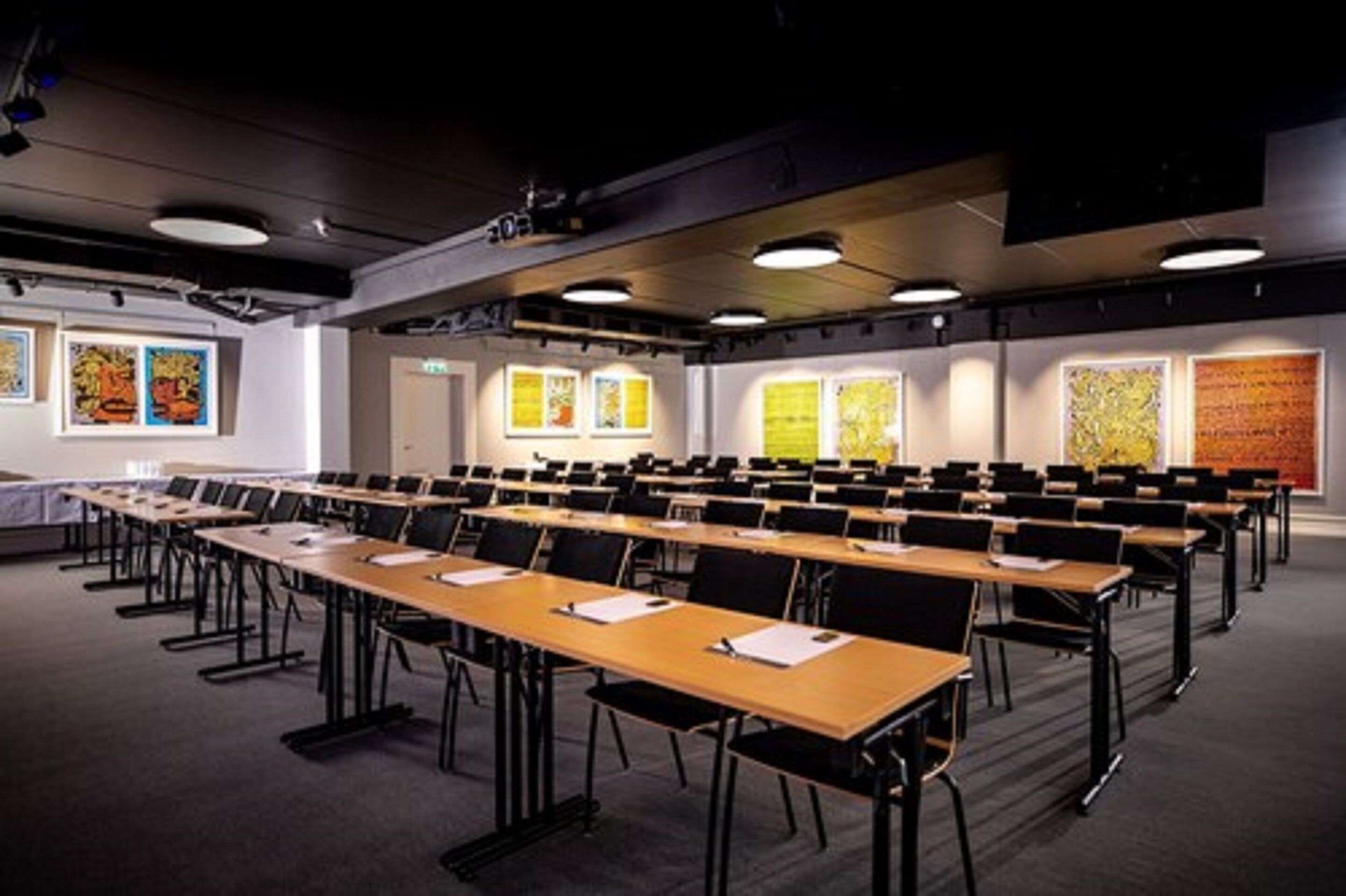 A conference room with rows of tables and chairs, large windows with artwork, and a dark ceiling with lighting fixtures.
