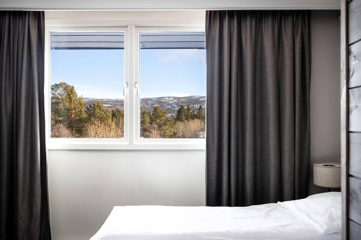 A hotel room with a bed, a window with curtains, and a view of trees and hills.