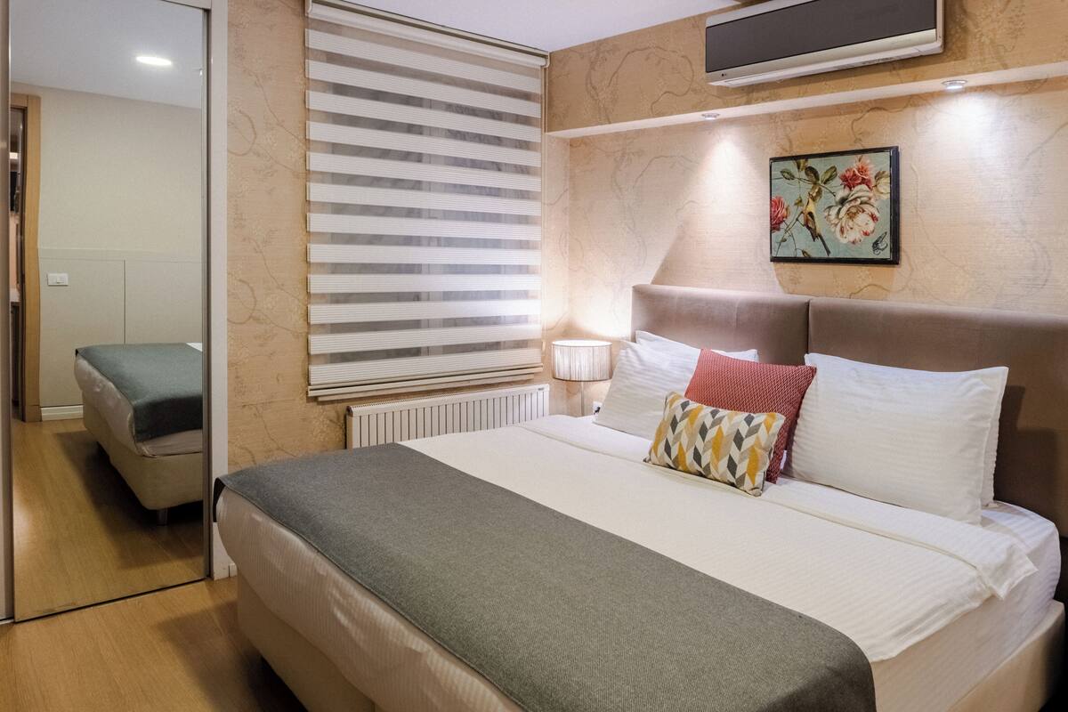 Una habitación de hotel con cama, una mesita de noche con lámpara, una ventana con persianas y un cuadro en la pared.