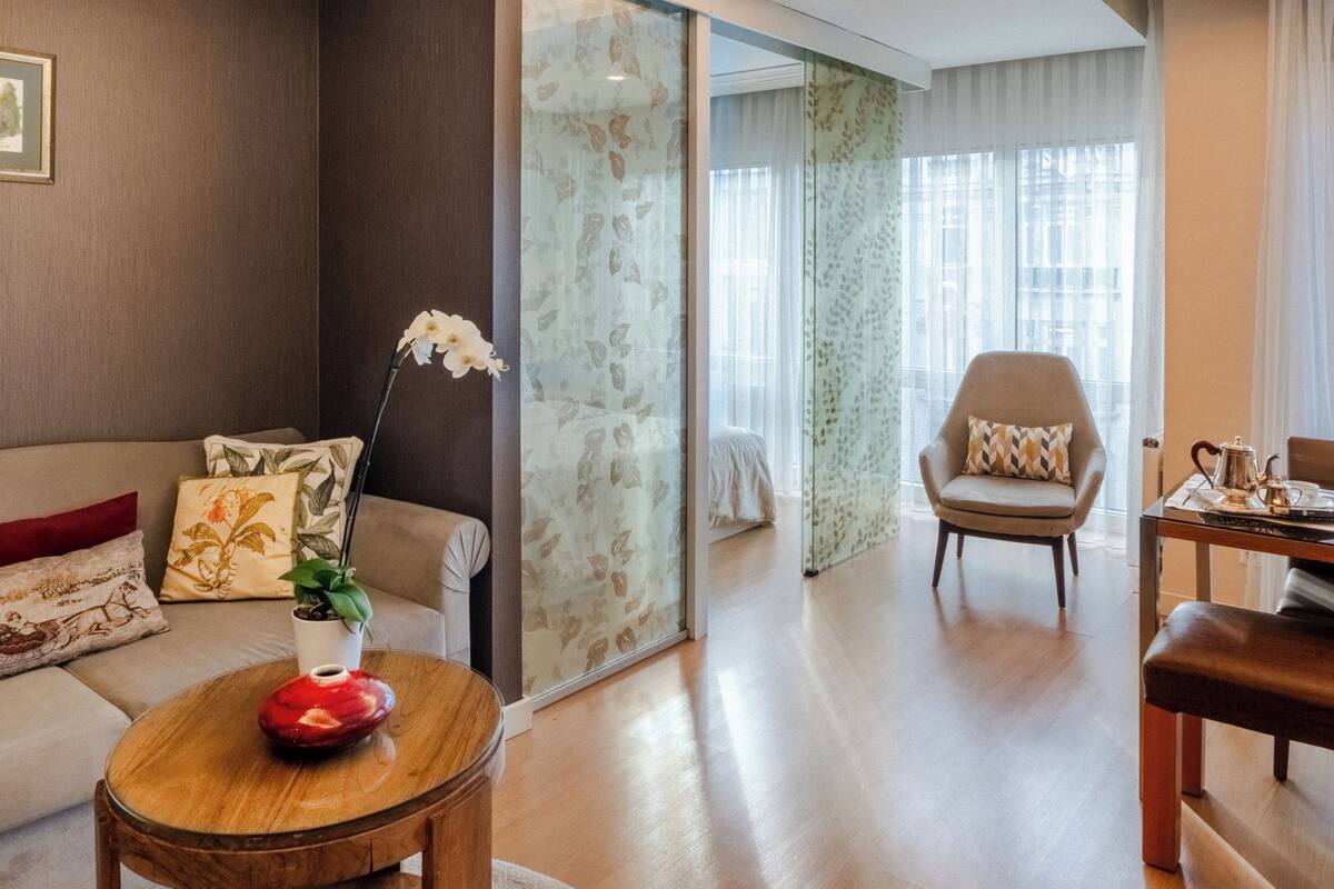 Una sala de estar moderna con un sofá, una mesa de centro y una silla. Hay una planta en la mesa de centro y un florero con una flor. La habitación tiene un ventanal con cortinas.