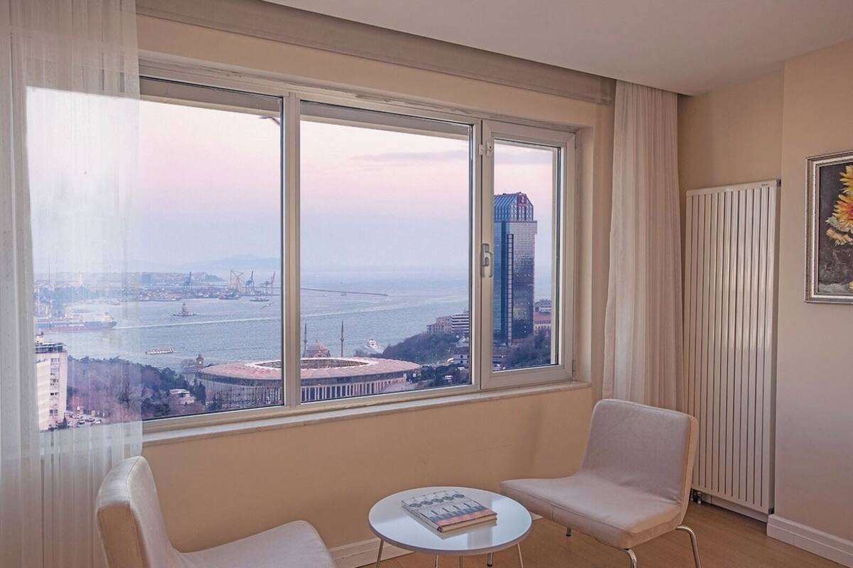 Habitación con un ventanal amplio que ofrece vistas a una ciudad y al agua. Hay dos sillas y una mesita con un libro.