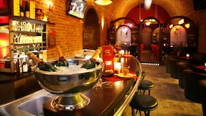 Una zona de bar con un bol grande de metal que contiene botellas de champán, un televisor montado en la pared y una pared de ladrillo con estantes llenos de botellas y copas.