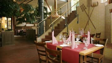 Un restaurante con paredes de ladrillo, mesas y sillas de madera. Se observa una escalera con barandilla y una planta colgando del techo.
