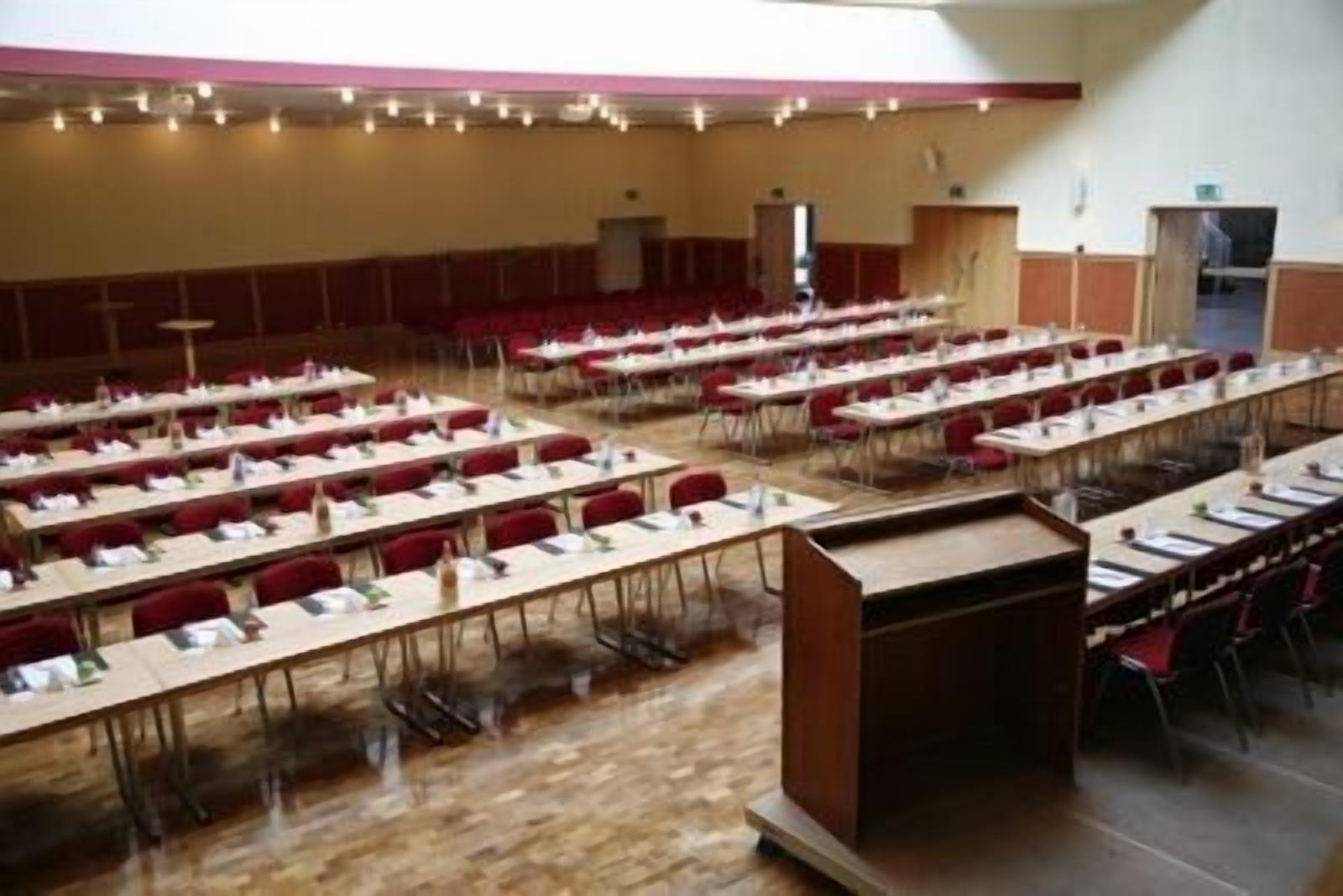 A conference room with multiple rows of tables and chairs, arranged in a U-shape, with a wooden floor and a high ceiling with lighting fixtures.
