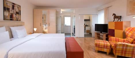 A hotel room with a large bed, a TV, a seating area with a plaid sofa, and a small kitchen in the background.