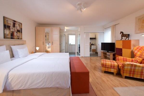 A hotel room with a large bed, a TV, a seating area with a plaid sofa, and a small kitchen in the background.