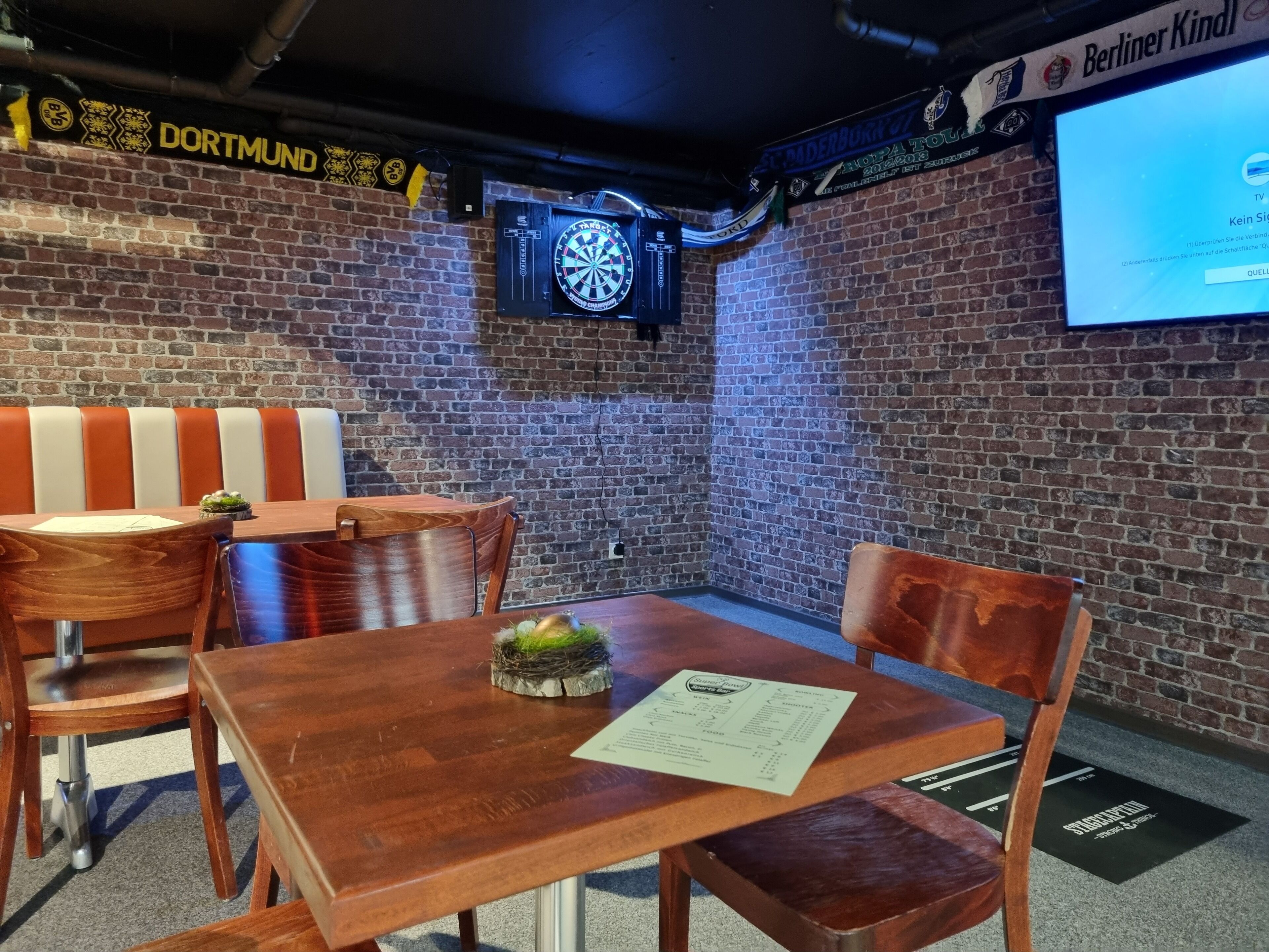 Une pièce avec un mur en briques, un jeu de fléchettes, un écran de télévision et une table avec des chaises.
