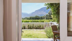 A view from a covered patio with a table and chair, overlooking a vineyard and mountains. - Auberge Clermont (Franschhoek)