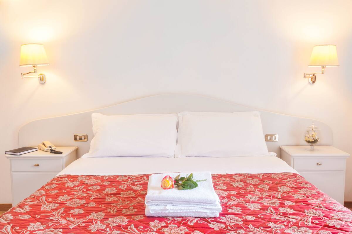 Una cama bien hecha con sábanas blancas y una colcha estampada de flores rojas, dos lámparas de noche, una mesita de noche con un teléfono y un libro, y un pequeño florero con flores en la mesita derecha.