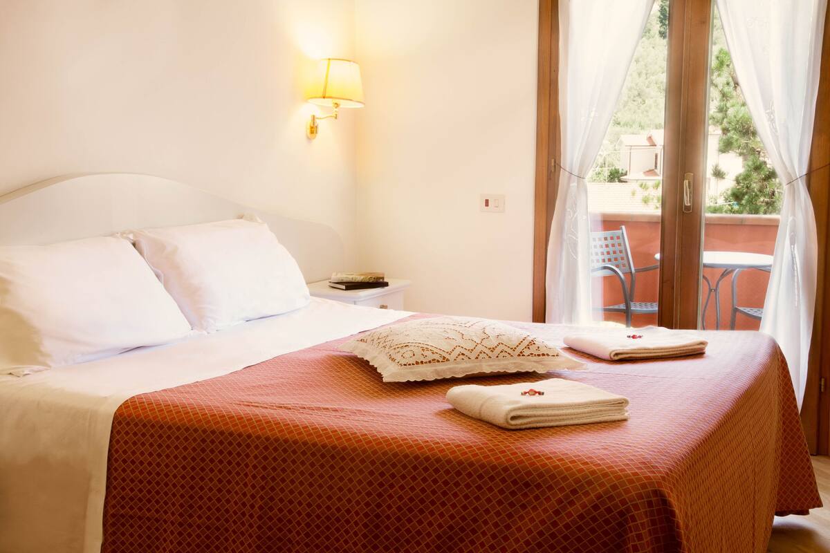 Habitación de hotel con cama, mesita de noche, lámpara y vistas al balcón exterior.
