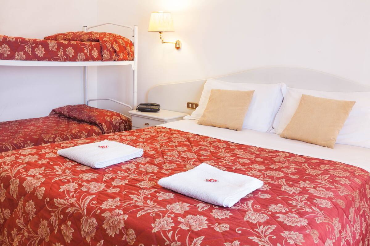 Habitación de hotel con literas, una cama individual y una mesita de noche con lámpara.