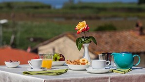 Mesa de café da manhã com vista para a água, um vaso branco com uma única rosa amarela e diversos itens de café da manhã, incluindo frutas, doces e uma chaleira azul.