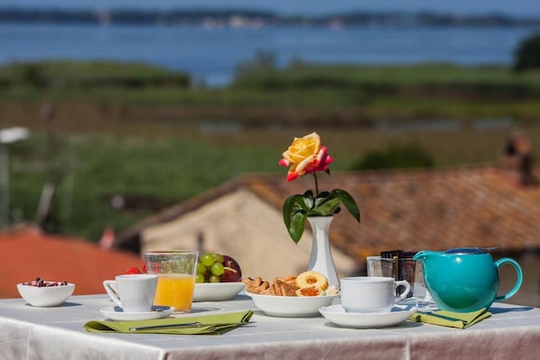 A breakfast table with a view of a body of water, a white vase with a single yellow rose, and various breakfast items including fruit, pastries, and a blue teapot. - Le Rotonde (Massarosa)