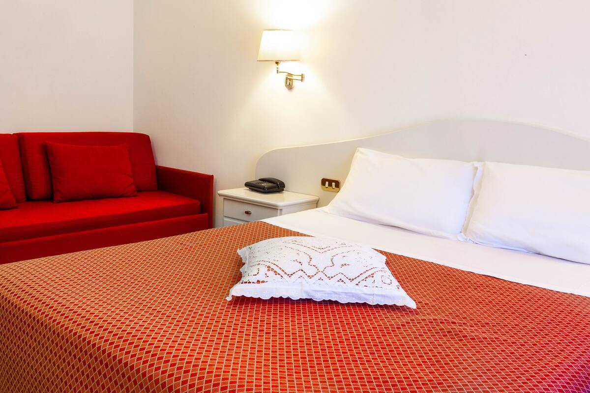 Una habitación de hotel con cama, sofá rojo, mesita de noche y lámpara.